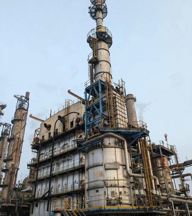 Detailed view of a large industrial oil refinery tower with intricate piping and structures against a clear blue sky.
